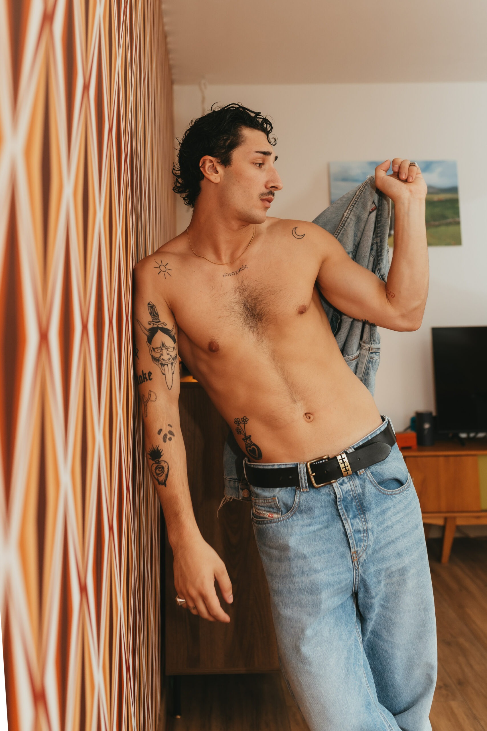 Théo en veste en jean, torse nu, appuyé contre le papier peint vintage